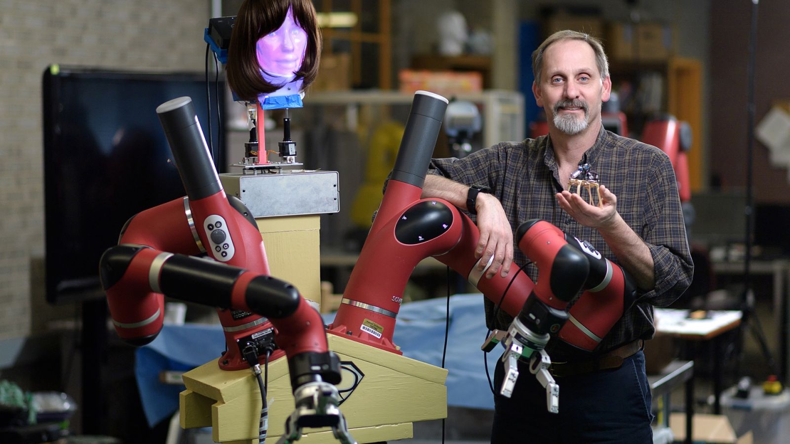 Richard Voyles stands with two examples of his research in robotics, Super Baxter (left) and NeUromorphic Computing on Laminated Electronic Organic substrate (NUCLEOs) in his hand. (Purdue University photo/John Underwood)